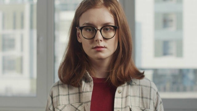 A young person with shoulder-length reddish hair and glasses looks directly at the camera.