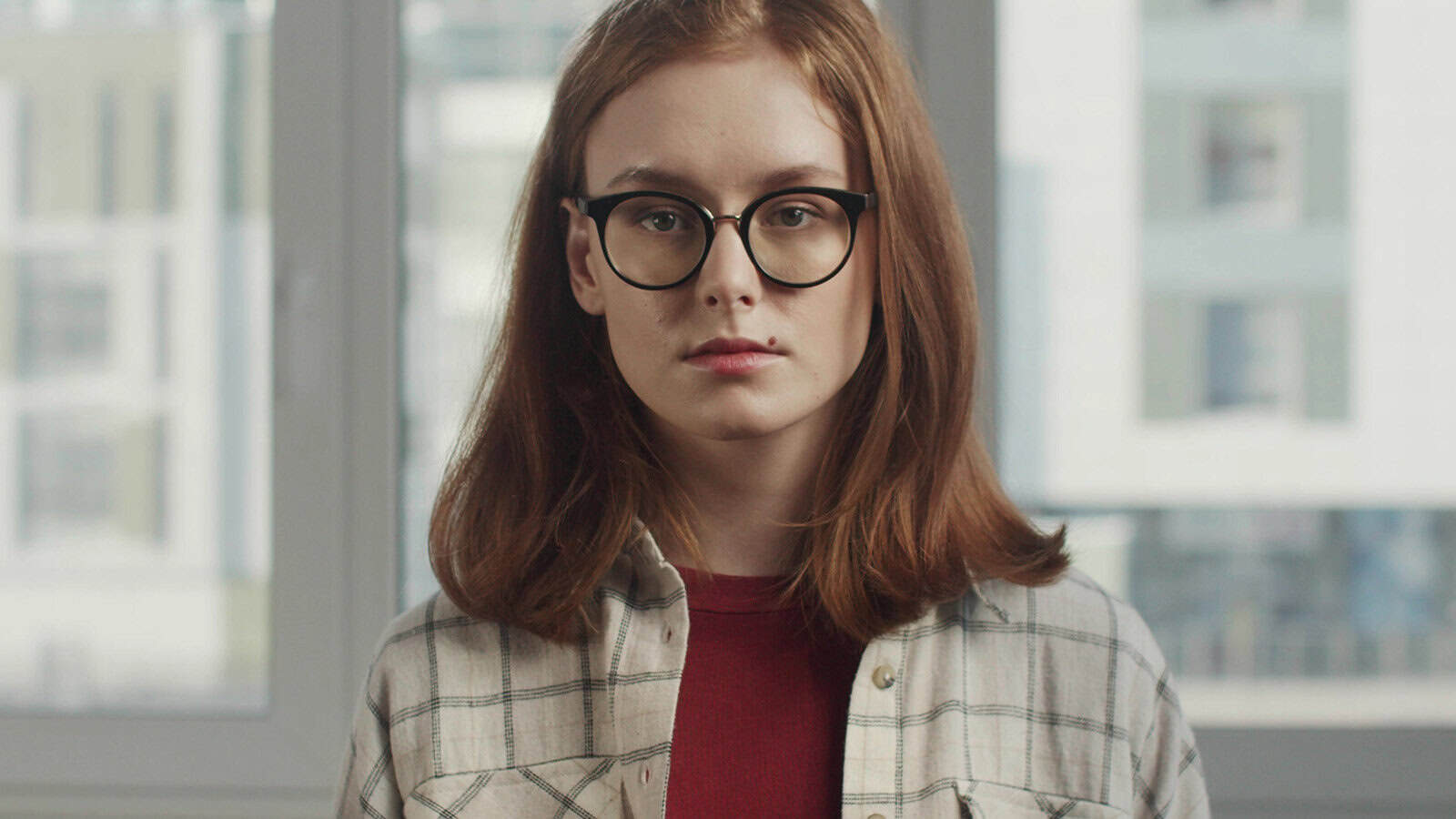 A young person with shoulder-length reddish hair and glasses looks directly at the camera.
