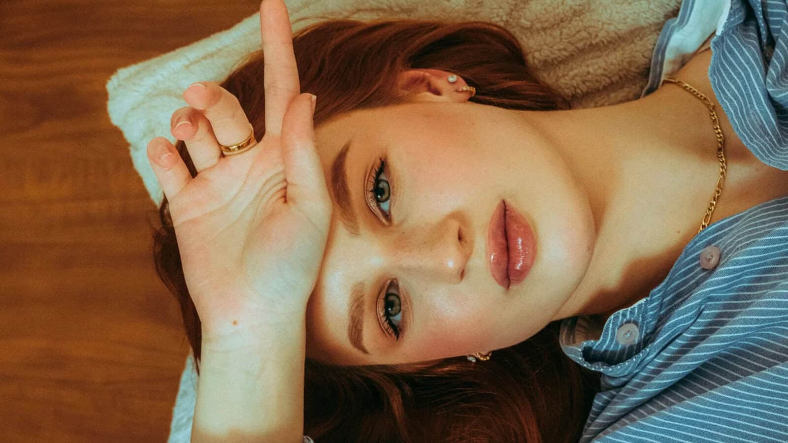 A close-up portrait of a person with auburn hair resting their hand against their forehead, wearing a blue pinstriped shirt and gold jewelry.
