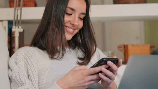 A woman with straight brown hair smiles while looking at her smartphone. She is sitting indoors, wearing a light sweater, and appears relaxed and content.