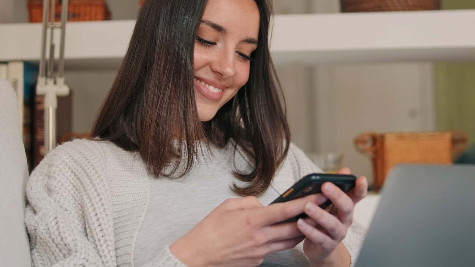 A woman with straight brown hair smiles while looking at her smartphone. She is sitting indoors, wearing a light sweater, and appears relaxed and content.