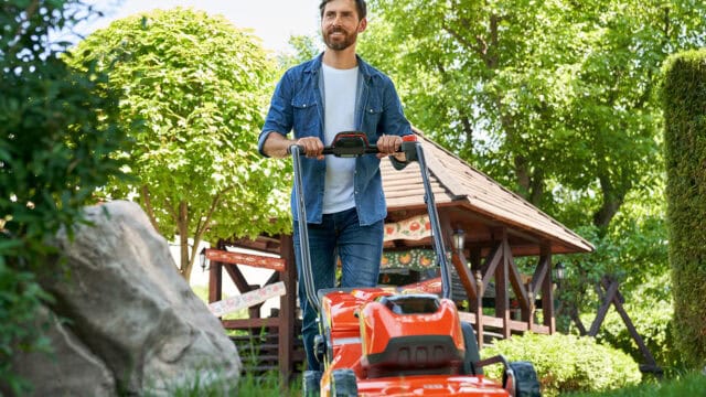 A smiling man pushing an orange lawnmower through a green garden.