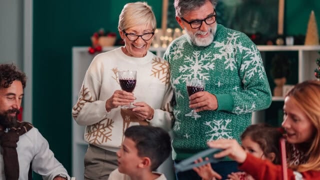 An older couple in Christmas sweaters stand smiling with glasses of red wine at a holiday gathering.