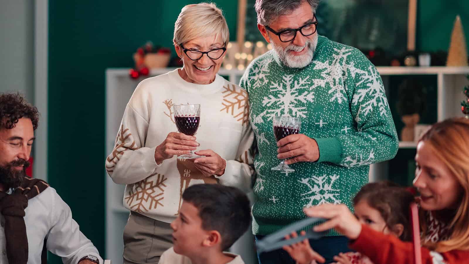 An older couple in Christmas sweaters stand smiling with glasses of red wine at a holiday gathering.