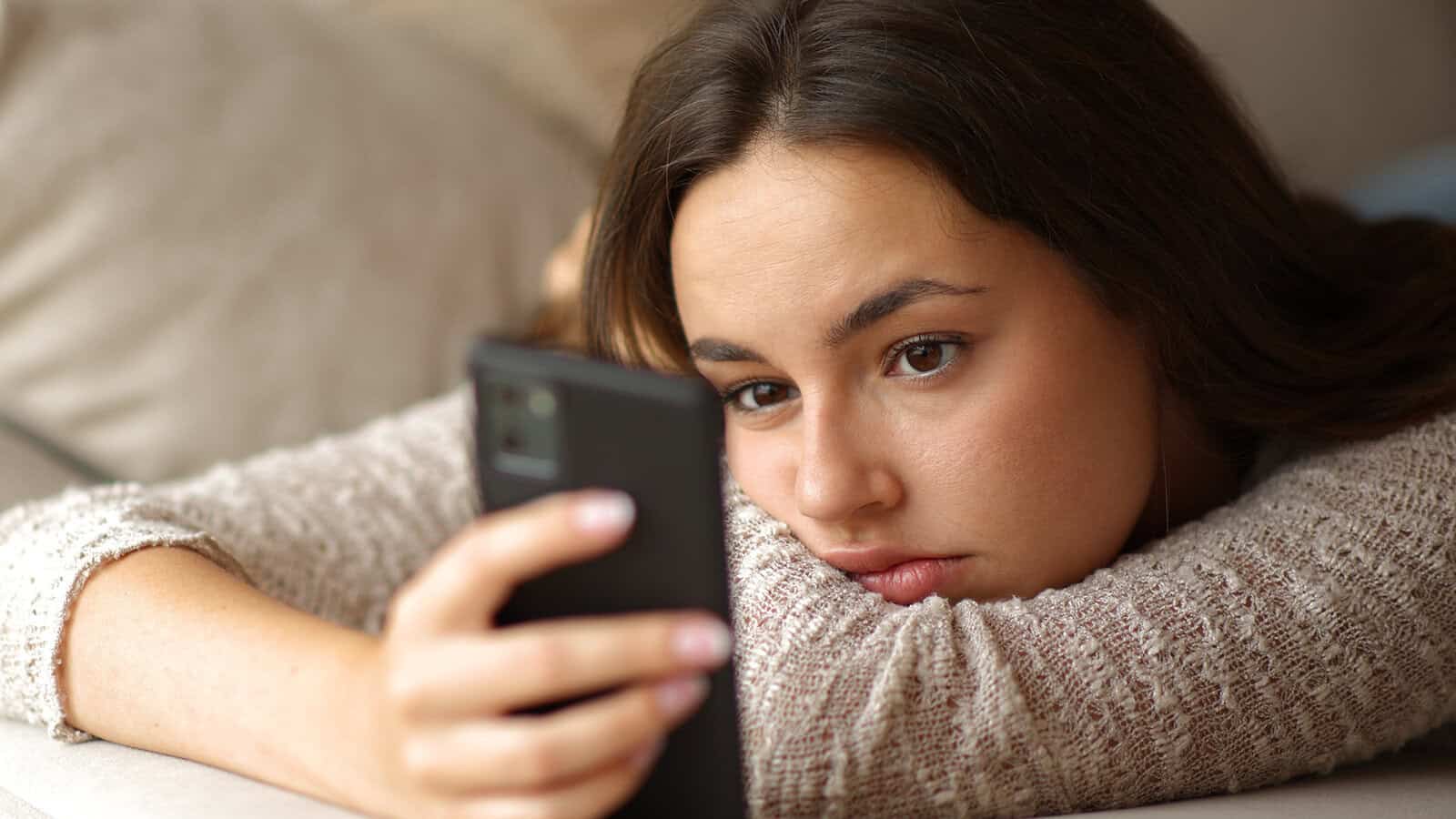A young woman lying down looks at her smartphone with a somber expression.