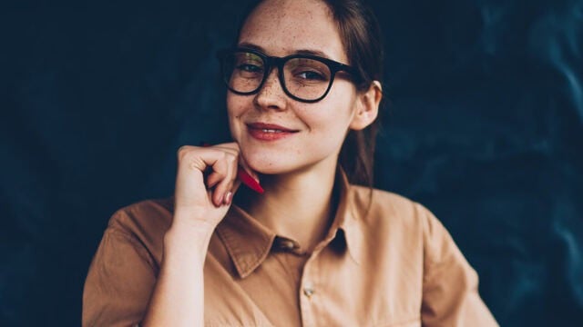 A woman wearing glasses rests her chin on her hand while holding a pen.