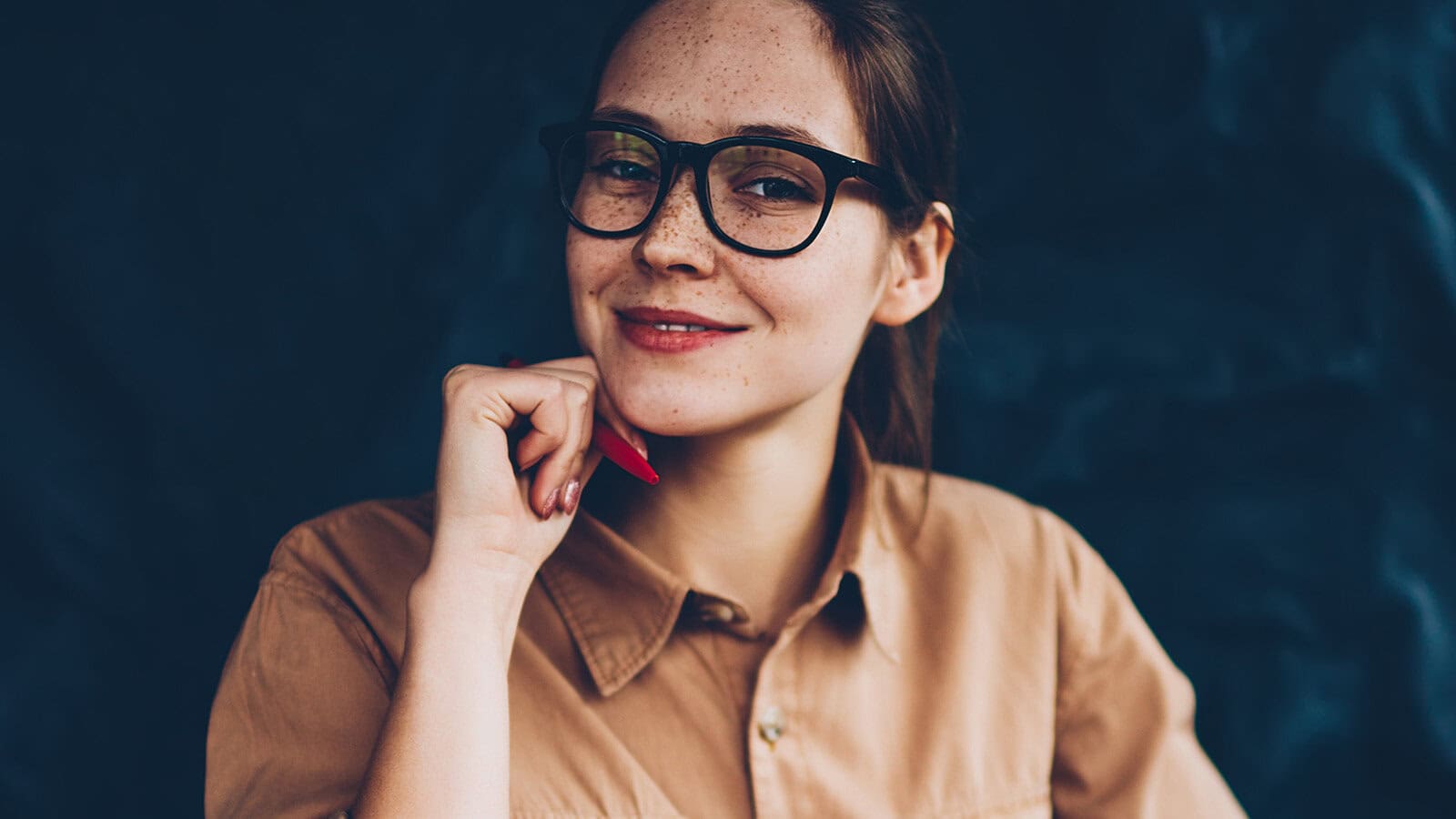 A woman wearing glasses rests her chin on her hand while holding a pen.
