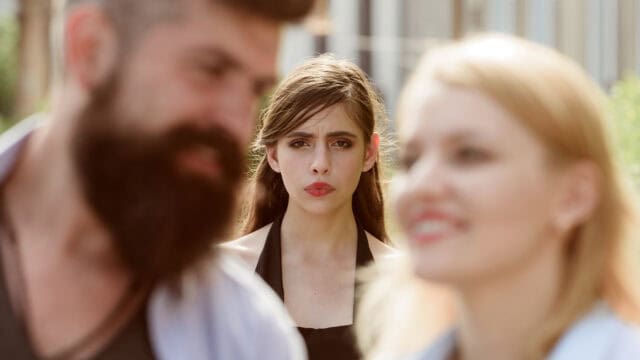 A man and woman in the foreground are out of focus, while a woman between them looks directly at the camera with a somber expression.
