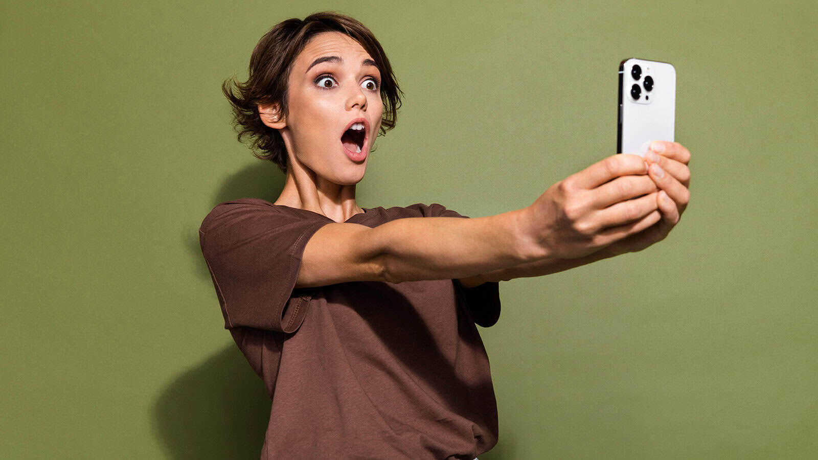 A woman with a surprised expression holding a smartphone to take a selfie.