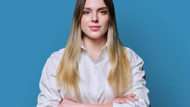 A woman in a white button-down shirt stands with her arms crossed against a blue background.