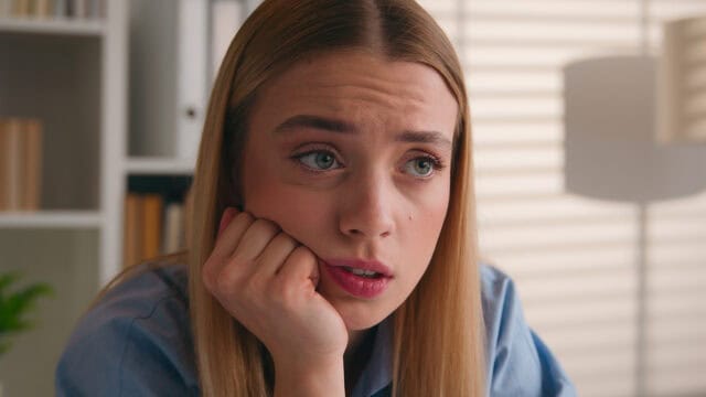 A young woman with a concerned expression rests her chin on her hand while looking off to the side.