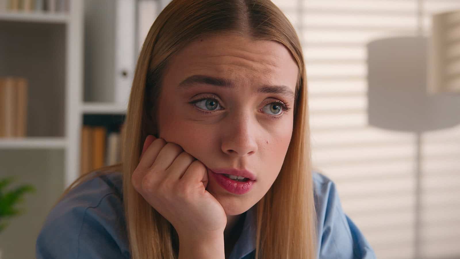 A young woman with a concerned expression rests her chin on her hand while looking off to the side.