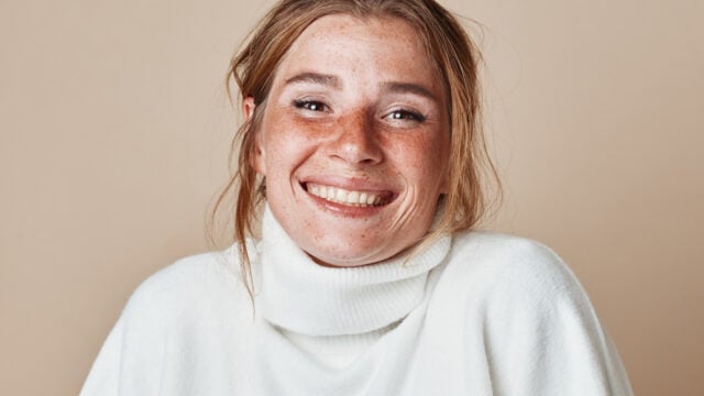 A smiling person with freckles wearing a white turtleneck sweater.