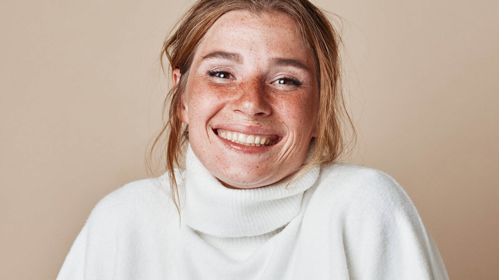 A smiling person with freckles wearing a white turtleneck sweater.