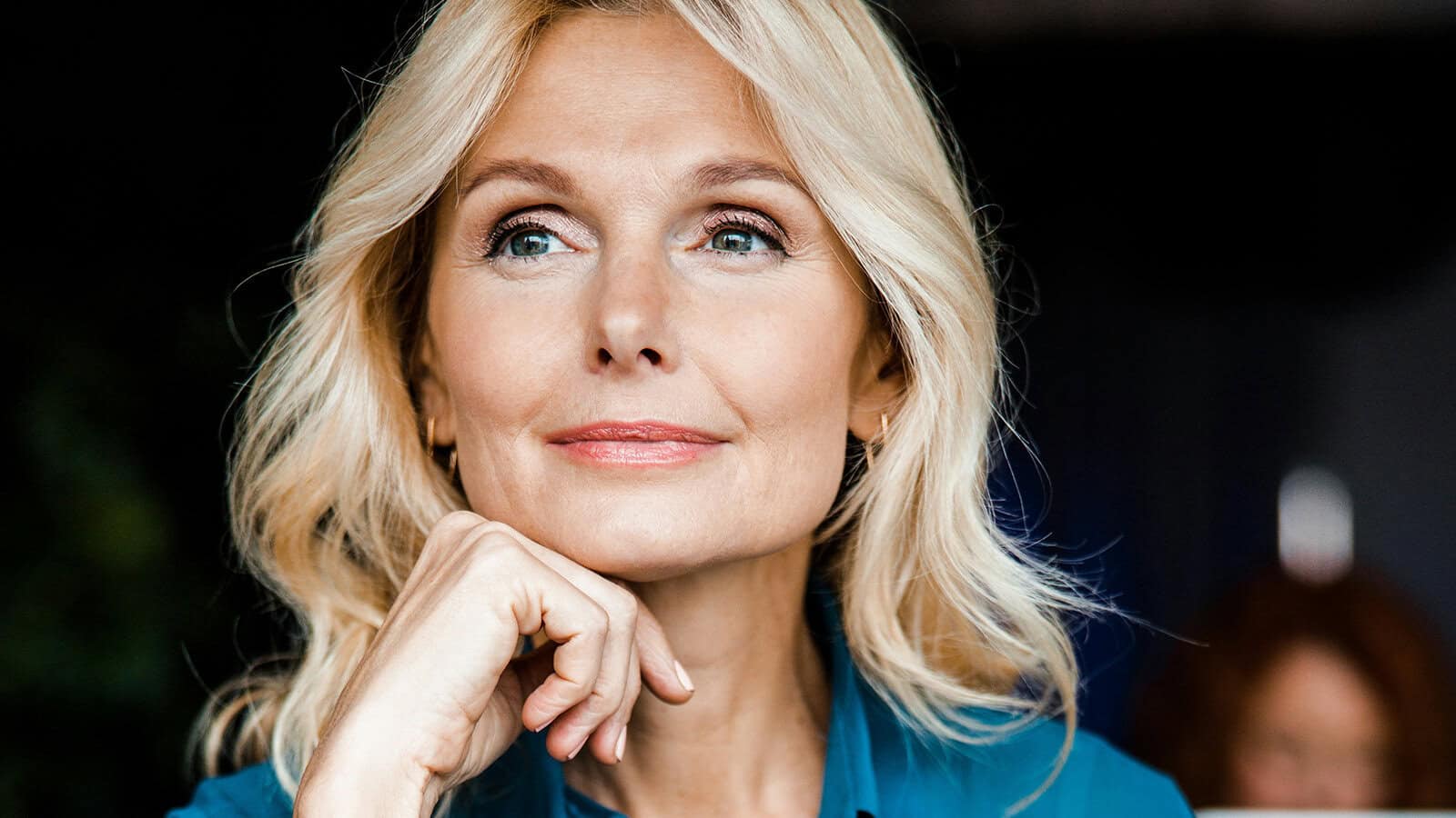 A blonde woman resting her chin on her hand, looking thoughtfully to the side.