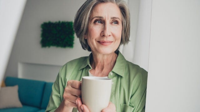 A smiling woman with gray hair wears a green button-down shirt and holds a white mug.