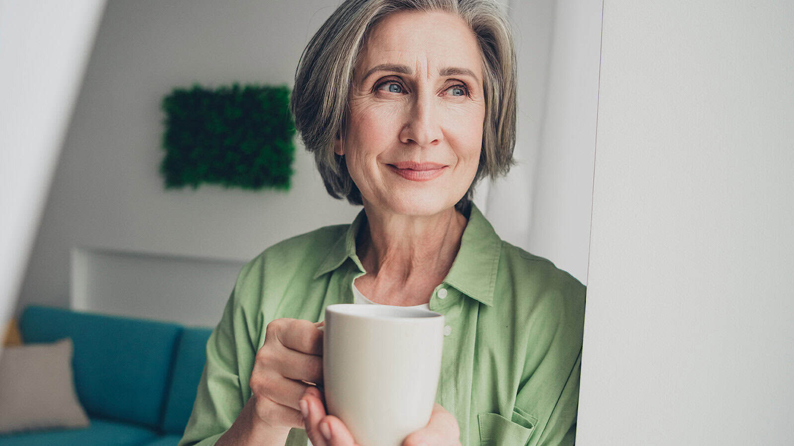 A smiling woman with gray hair wears a green button-down shirt and holds a white mug.