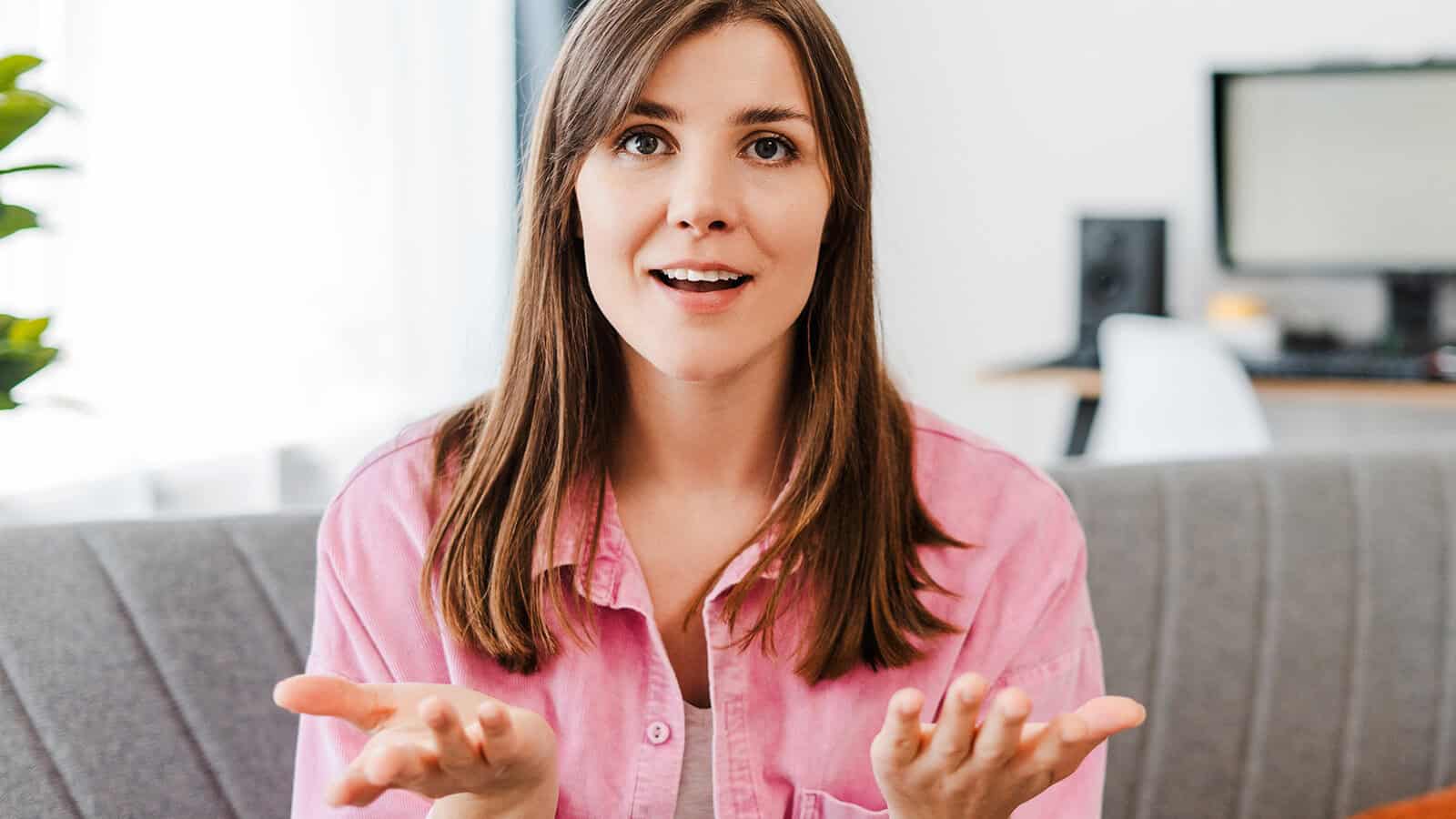 A woman in a pink shirt sits on a sofa, speaking and gesturing with her hands.