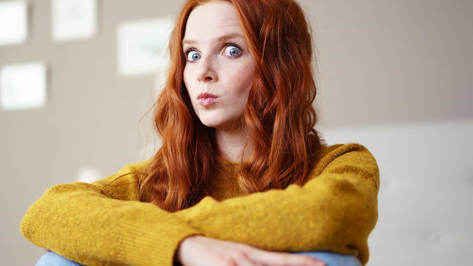A woman with long red hair wearing a mustard yellow sweater makes a surprised expression with her lips pursed.