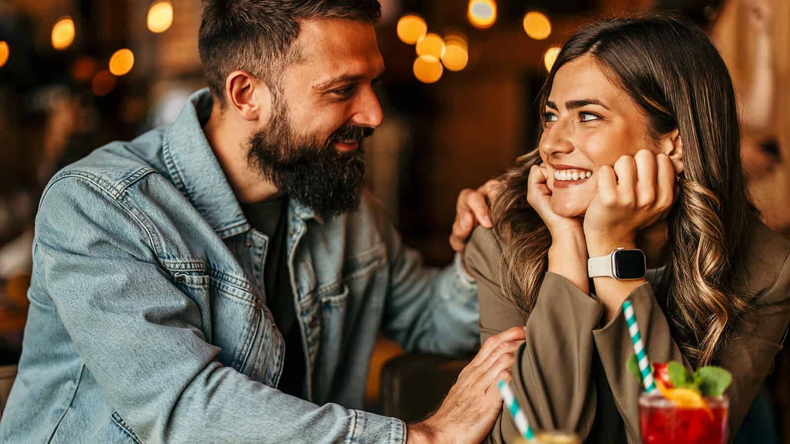 A man with a beard looks affectionately at a smiling woman while resting his hand on her shoulder at a restaurant.