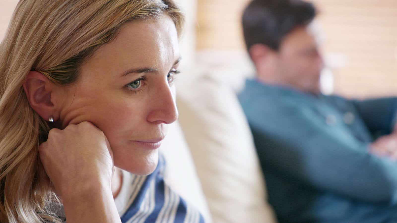 A concerned woman resting her chin on her hand while a man sits out of focus behind her.