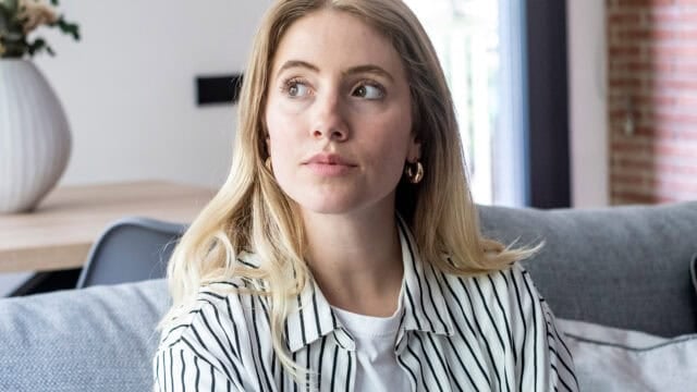 A woman with blonde hair wearing a striped shirt and gold hoop earrings sits on a sofa, looking off to the side.