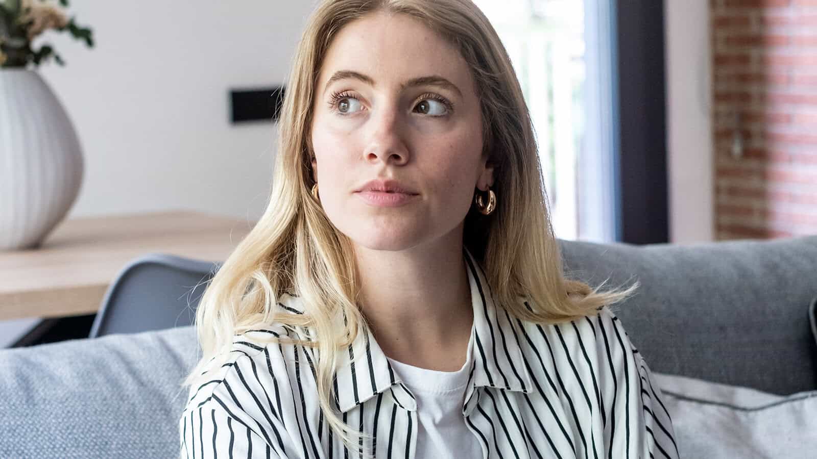 A woman with blonde hair wearing a striped shirt and gold hoop earrings sits on a sofa, looking off to the side.
