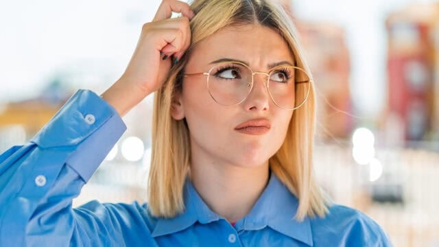 A woman with blonde hair and round glasses scratches her head with a thoughtful or confused expression.