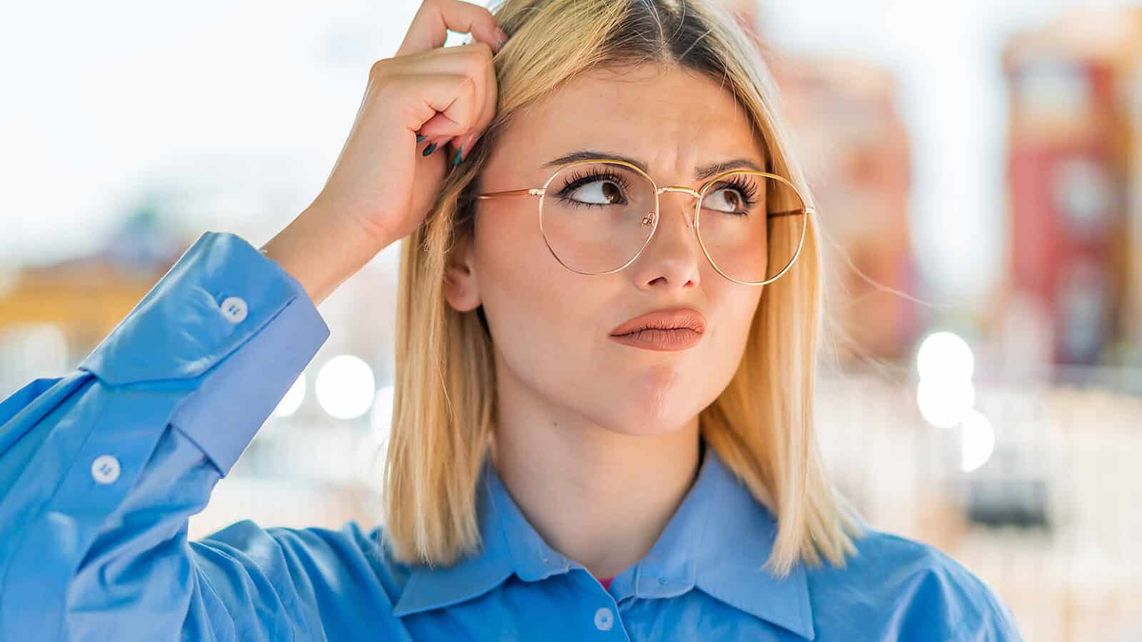 A woman with blonde hair and round glasses scratches her head with a thoughtful or confused expression.