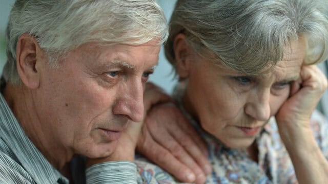 An elderly man rests his hand on the shoulder of an elderly woman as they look down with sad expressions, conveying the heavy emotional toll of navigating aging parents.