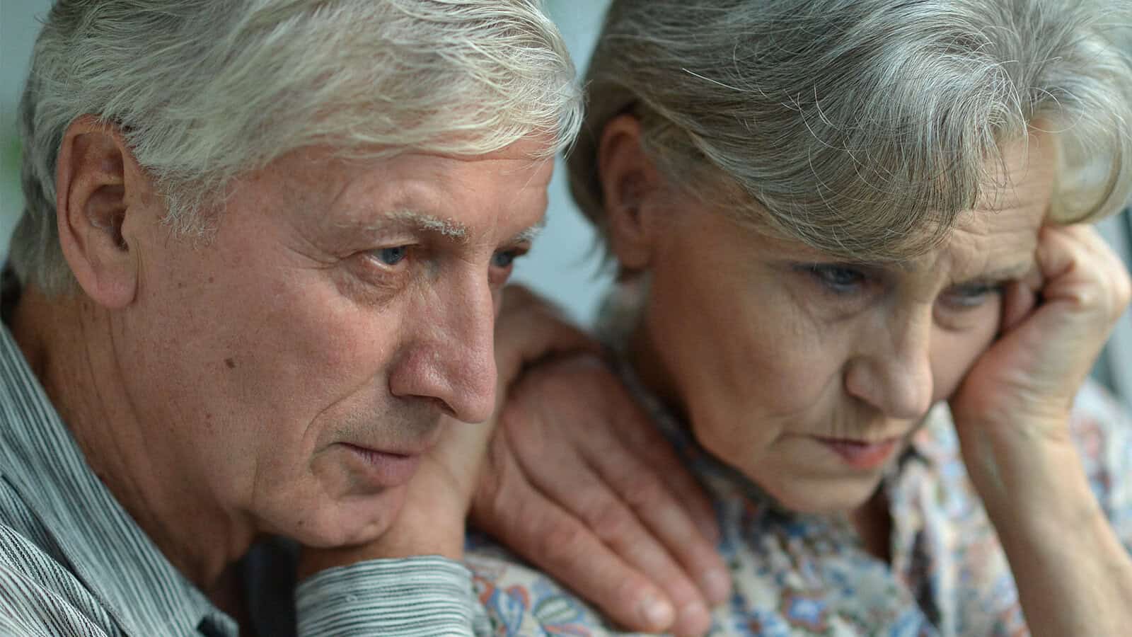 An elderly man rests his hand on the shoulder of an elderly woman as they look down with sad expressions, conveying the heavy emotional toll of navigating aging parents.