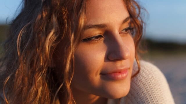 A young woman with wavy brown hair smiles softly, looking to the side. She is illuminated by warm sunlight and wears a light-colored sweater—her expression reflecting quiet resilience. The background is softly blurred.