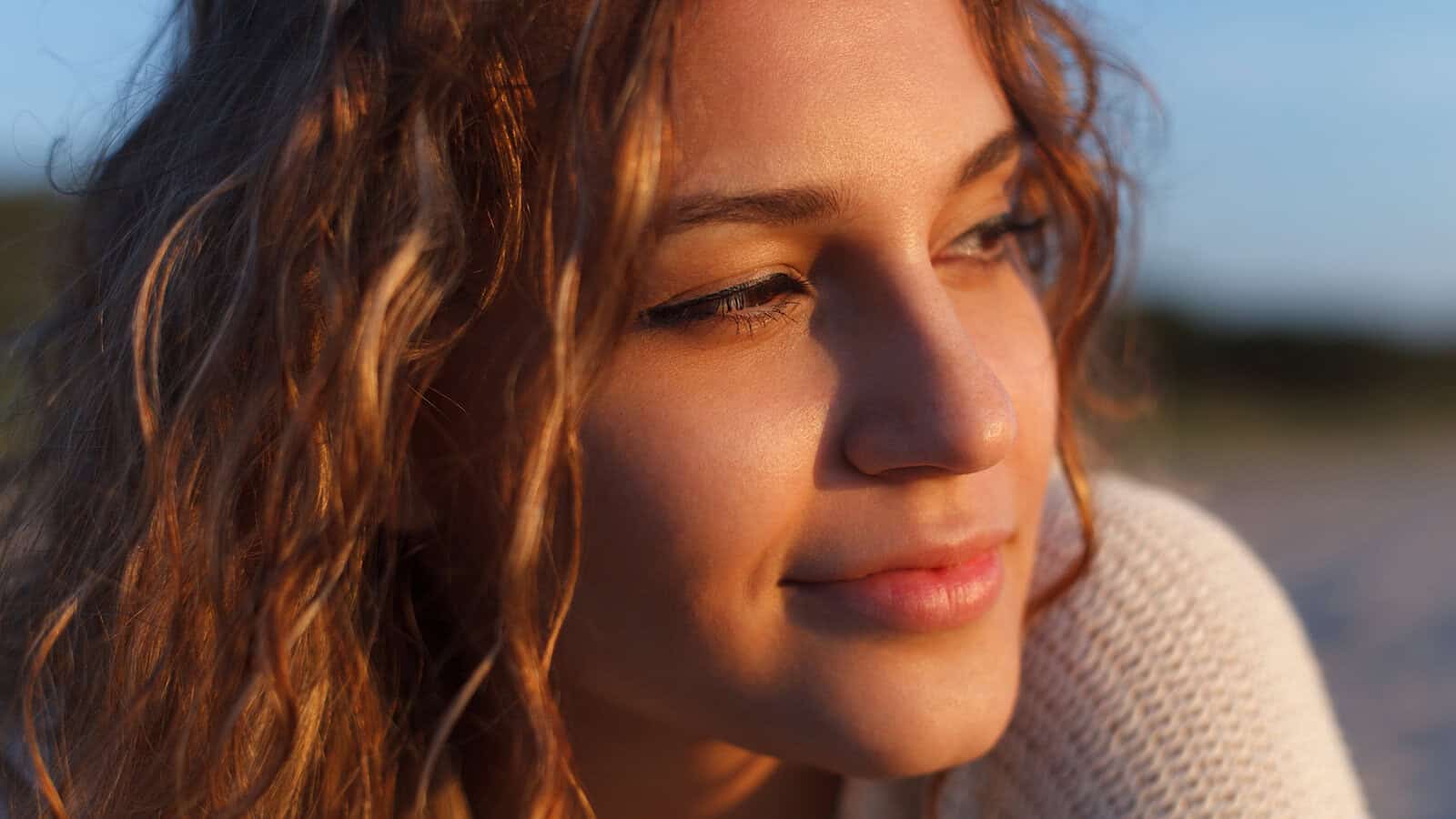 A young woman with wavy brown hair smiles softly, looking to the side. She is illuminated by warm sunlight and wears a light-colored sweater—her expression reflecting quiet resilience. The background is softly blurred.