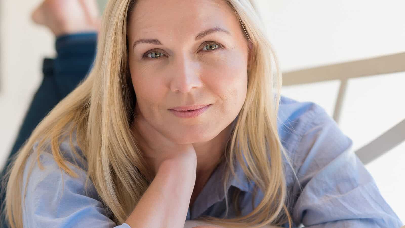 A woman with long blonde hair and light eyes, wearing a light blue shirt, rests her chin on her hand and looks directly at the camera with a gentle expression, reflecting quietly on caring what others think.