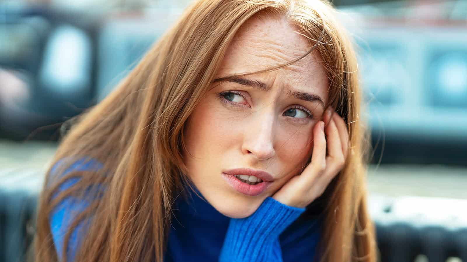 A woman with long red hair in a blue sweater rests her face on her hand, looking worried or concerned, perhaps reflecting on the strength it takes to break old habits, with a blurred outdoor background.