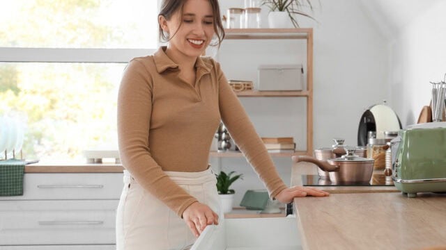 A smiling woman in a tan long-sleeved shirt opens a kitchen drawer, showcasing a calming personality pattern for kitchen organization.