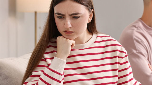 A woman with long brown hair and a red-and-white striped sweater sits indoors, resting her chin on her hand and looking down with a concerned or thoughtful expression, perhaps reflecting on adjusting friendships or the experience of losing friends.