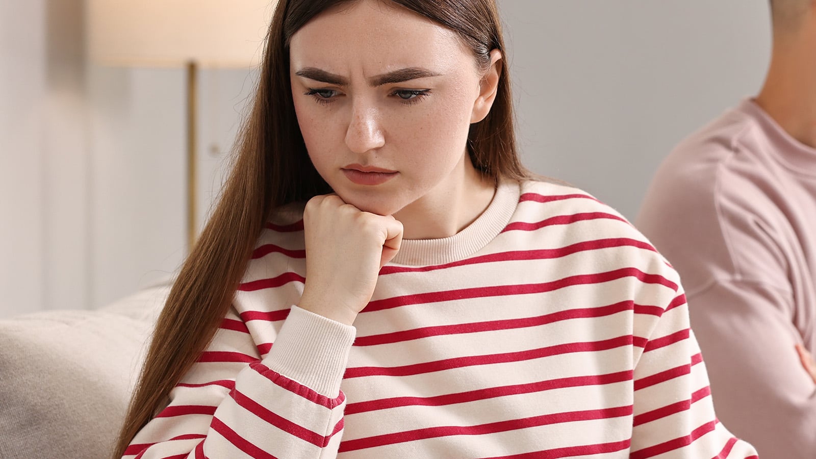 A woman with long brown hair and a red-and-white striped sweater sits indoors, resting her chin on her hand and looking down with a concerned or thoughtful expression, perhaps reflecting on adjusting friendships or the experience of losing friends.