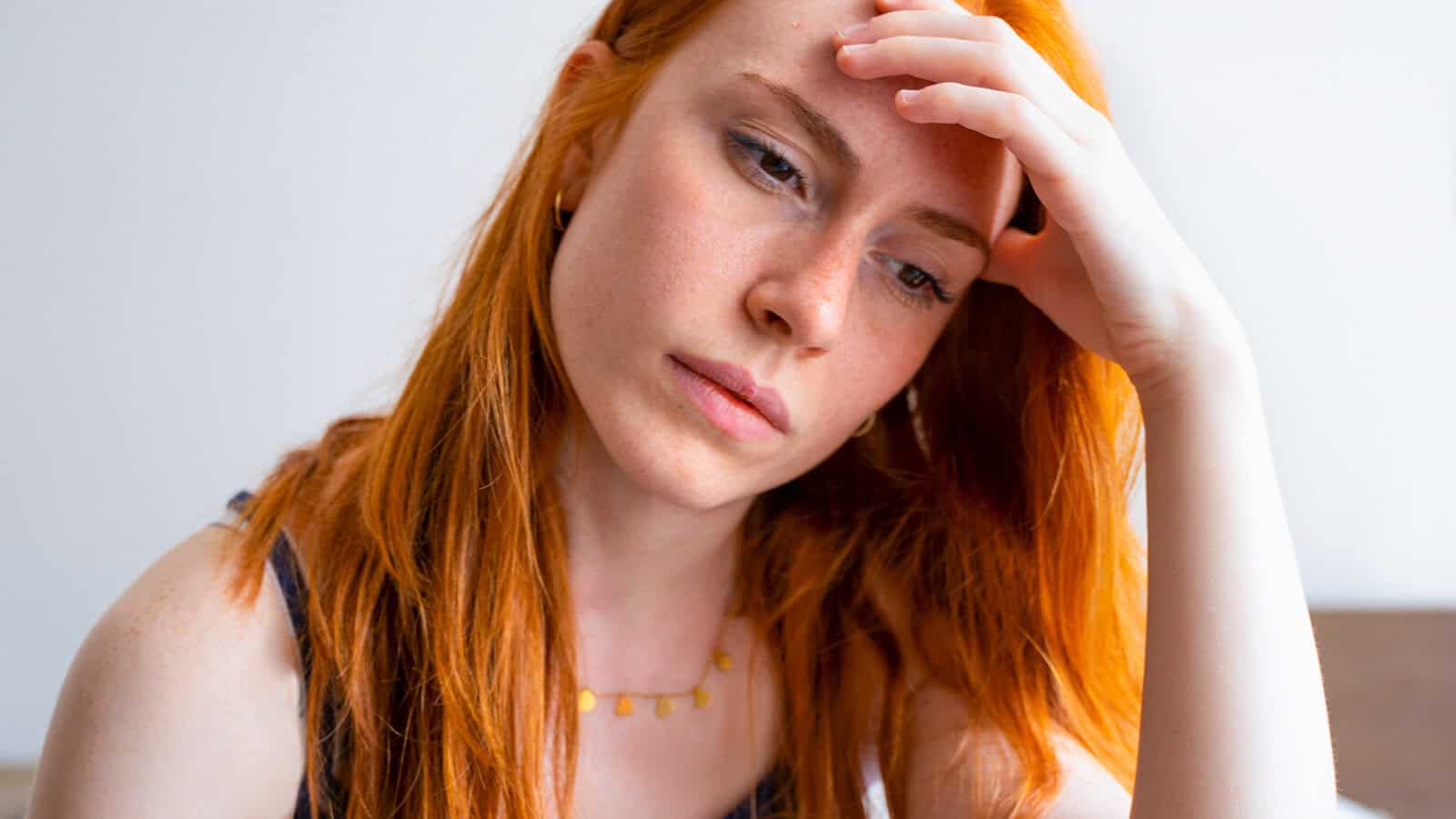 A woman with long red hair rests her forehead on her hand, looking down with a thoughtful or worried expression, as if reflecting on approval patterns and how to say no without feeling guilty.
