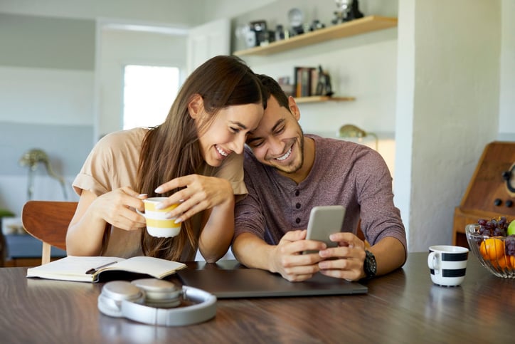 Cheerful boyfriend showing mobile phone to girlfriend in dining room. Young woman is holding coffee cup. They are spending leisure time together at home.