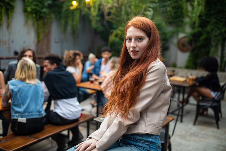 Young Charming charismatic Ginger woman sitting in pub, seriously looking at camera, friends sitting behind her