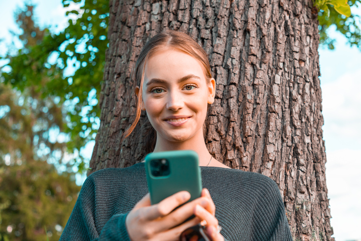 Happy young woman in the garden reading messages on her mobile phone.