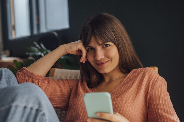 Beautiful cheerful young Caucasian woman wearing a pink blouse and jeans looking at camera using a mobile phone while sitting casually in a chair.