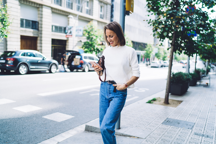 Happy lady in casual outfit using mobile on street while walking hand in pocket on modern city street on sunny day