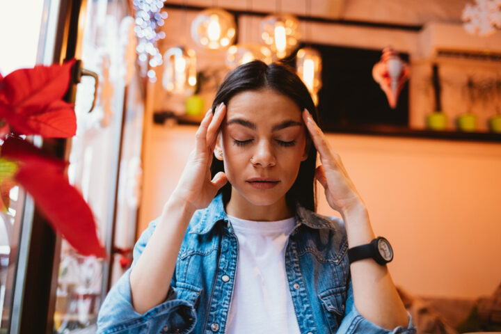 Portrait of a young tired woman in a denim jacket holds her head with both hands in a cafe