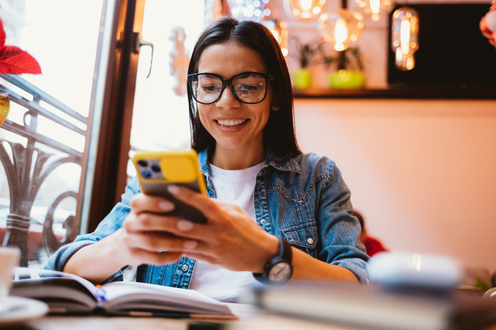 Happy young woman student holding smart phone over her textbook sitting at table in cafe.