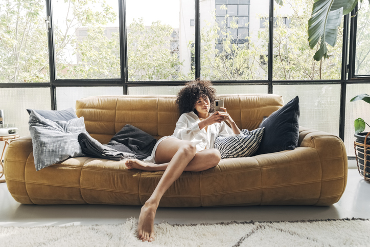Pretty Young African American woman with curly hair sitting on comfortable sofa texting a friend. Home concept. Technology concept.