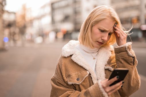 Distraught and worried young woman using phone on city street. She is texting or reading bad news.