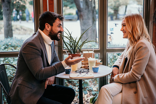 Spontaneous photo of a cute and cheerful young adult couple in love, sitting in their favorite restaurant and enjoying their talk, both elegantly dressed. A handsome bearded guy is talking to his cute blonde with excitement and joy