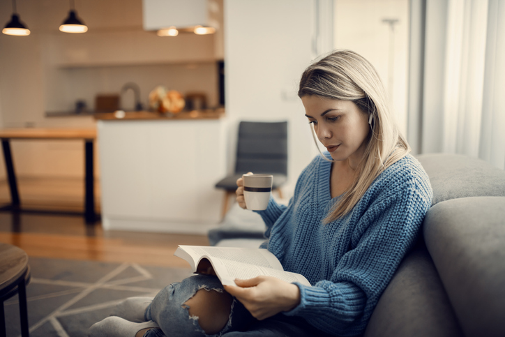 A focused woman reading a book and drinking coffee at her cozy home.